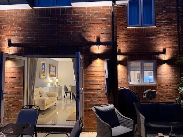 Brick rear elevation of a house at night lit by a row of black up and down wall lights above a patio seating area